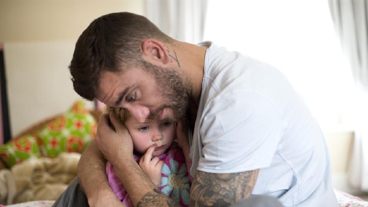 A father comforts his upset little girl, he hugs her tightly. The little girl looks upset and tired.