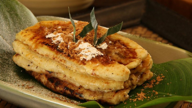 Pisang Plenet, which means flattened bananas, are indigenous street food snack from Semarang, Central Java. These plantain bananas are grilled, flattened, then topped with palm sugar and caster sugar. A serving contains three to four bananas; and it is served on a ceramic plate that has been lined with small banana leaf.  The plate is placed on a woven bamboo tray. In the background is a bowl of palm sugar.