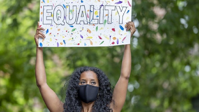 A beautiful young female adult of African ethnicity is standing outdoors. She is wearing a face mask and is holding a poster with the word 