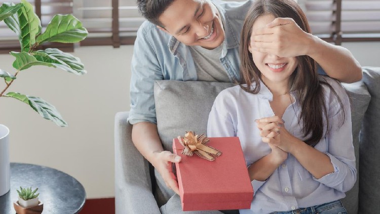 Handsome man is giving his girlfriend a red gift box for birthday surprising in the living room at home