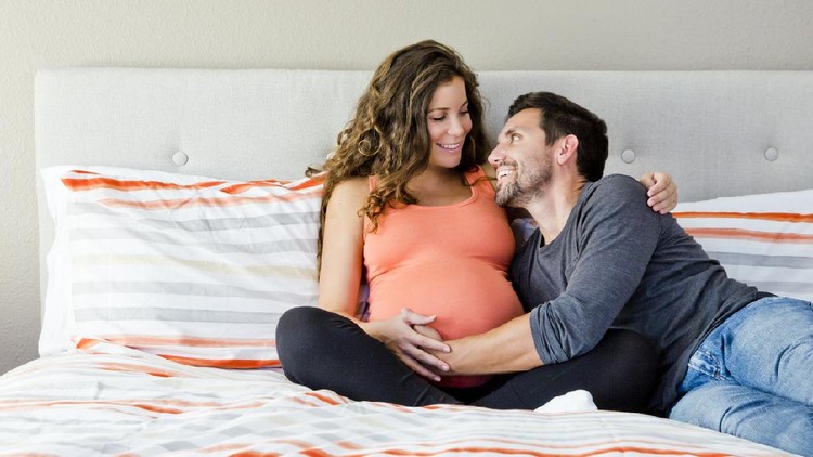 Young happy pregnant couple sitting on a bed and smiling. Man looking at woman. Lifestyle maternity photo.