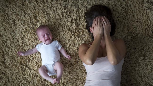 Ibu Mengalami Baby Blues - Seorang ibu baru yang terlihat cemas dan lelah, duduk di sofa dengan bayinya, menggambarkan perasaan emosional yang umum setelah melahirkan