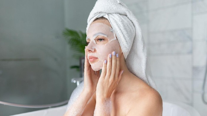 Beautiful young woman sitting in bathtub with sheet mask on face. Close up portrait of pretty girl in bath towel on head and doing beauty treatment. Home bathroom interior. Spa procedure. Skincare