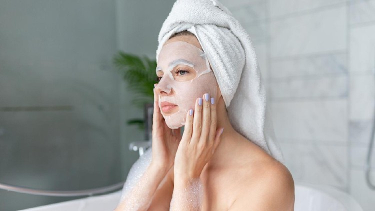 Beautiful young woman sitting in bathtub with sheet mask on face. Close up portrait of pretty girl in bath towel on head and doing beauty treatment. Home bathroom interior. Spa procedure. Skincare