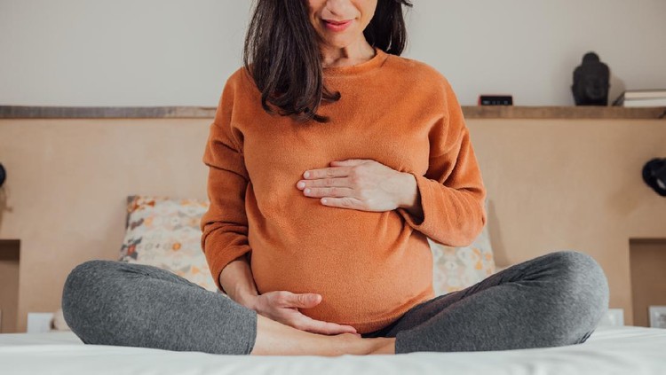 Front view of cropped long hair brunet caucasian pregnant woman, sitting in crossed legs, in yoga pose, in bed at home, holding her belly with hand. Maternity, pregnancy, new life.