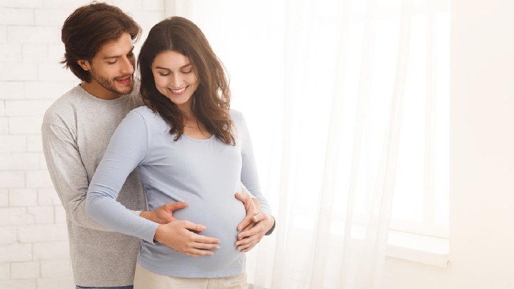 Loving man hugging his pregnant wife from behind standing near window at home, copy space