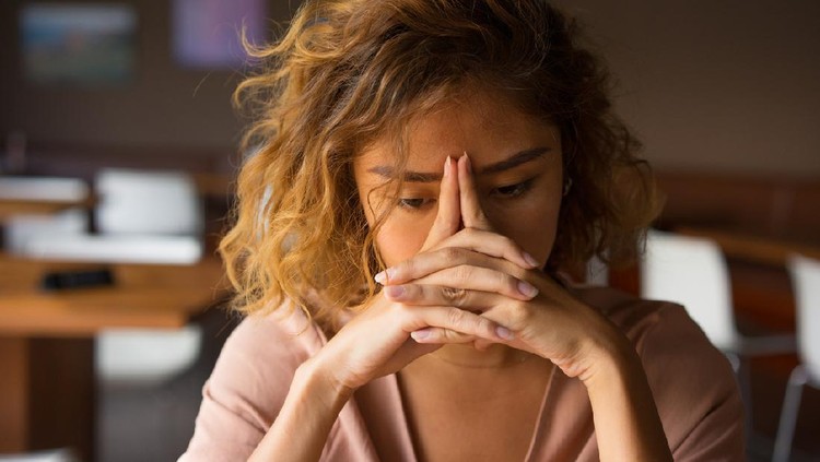 Closeup of sad young Asian woman at cafe leaning head on clasped hands and staring into vacancy. Tired freelancer feeling burnout. Stress and bad news concept