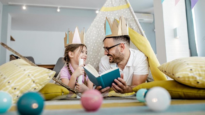 Young Asian mother sitting on the floor in the bedroom reading book to little daughter, enjoying family bonding time together at home
