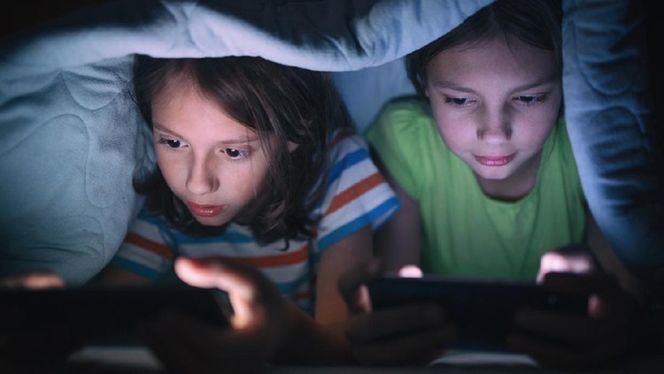 Boy and girl playing games on mobile phones while lying on bed in bedroom under the blanket. They are spending some nice time together that makes them happy.
