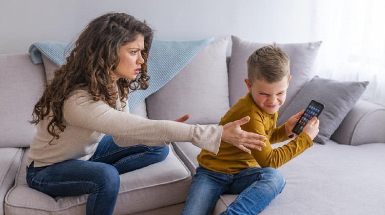 Boy having a tantrum and fighting with his mother for a smart phone sitting on a couch in the living room at home. Boy plays with electronic device but his mother not happy with this
