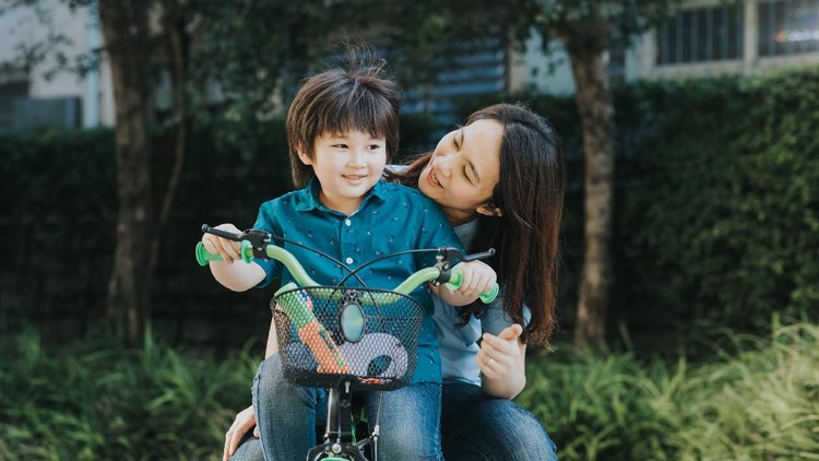 Asian mother and son riding a bicycle together outdoors in a city park.