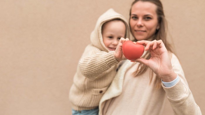 Mom woman with a little boy son 3-5 years old, background wall. In the hands of a toy red heart, concept give life and love, donation. Mother and St. Valentine's Day. Free space for text.