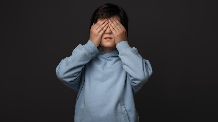 Portrait of boy 10-12 years old, dressed in casual clothes covering his eyes with hands. Human emotions concept. Studio shot, white background