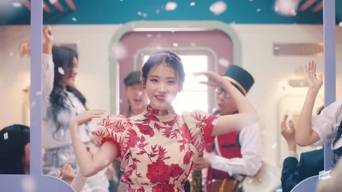 Kembali di suasana gerbong kereta, IU terlihat menari bersama dengan para penumpang. IU memakai dress bermotif floral kombinasi warna merah dan krem. Seyuman manisnya tampak menyempurnakan scene ini. (Foto:youtube.com/1theK)