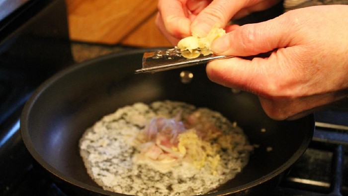 Close-up of a woman's hands grating garlic into a frying pan with butter, red onion, and shallots.