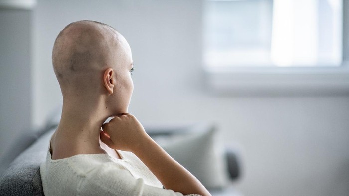 A woman with a shaved head is indoors in her house. She is sitting on her couch and looking into the distance.