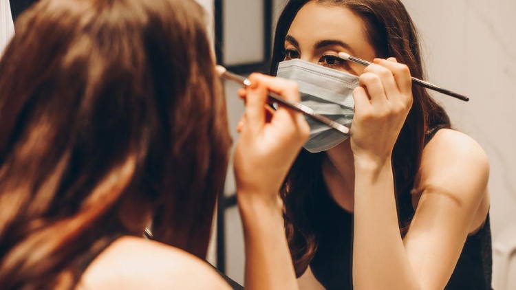 1253160478 selective focus of young woman in medical mask and black dress applying eye shadow in bathroom