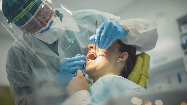 The dentist, fully dressed in a protective suit with an eyewear protection is with a patient. A woman in the chair has her mouth opened and examined while holding a suction pump.