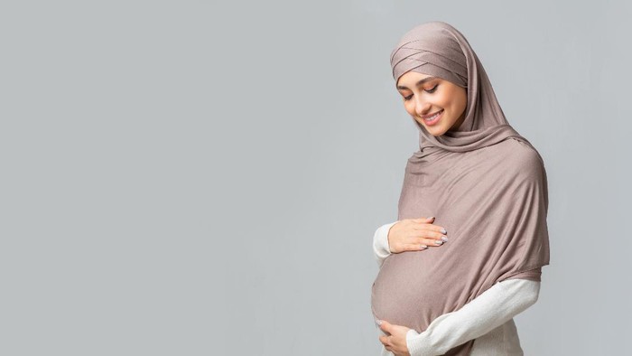 Joyful Pregnant Muslim Lady Using Digital Tablet Browsing Internet Sitting On Couch At Home. Pregnancy Lifestyle Concept. Arab Mother-To-Be Using Computer. Copy Space For Text
