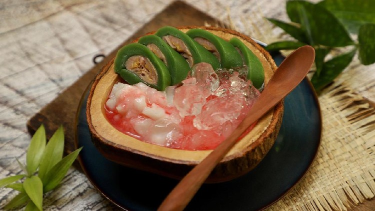 Es Pisang Ijo, the popular dessert of plantain banana roll with coconut milk custard and red sugar syrup from Makassar (Ujung Pandang), South Sulawesi. The banana is wrapped with pandan crêpe; then served in slices on the custard and drenched with red sugar syrup. The dessert is plated on a wooden bowl and placed on ceramic plate. A wooden spoon is placed on the rim of the bowl. Several sprigs of bamboo leaves are used for table decoration.