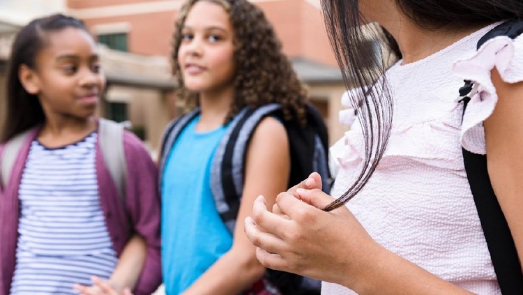 Ilustrasi Anak Antisosial Two schoolgirls talk with each other, ignoring a lonely classmate. They are standing outdoors on campus.