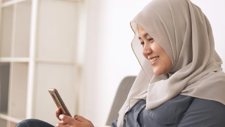 Asian muslim woman sitting of the sofa at home using phone and smiling. Feeling happy. Female doing communication on her smart phone, sending message or browsing online