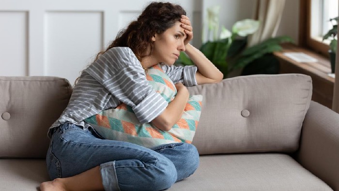 Frustrated young lady sitting on sofa, cuddling pillow, looking away at window. Lost in thoughts unhappy stressed millennial woman regretting of wrong decision, spending time alone in living room.