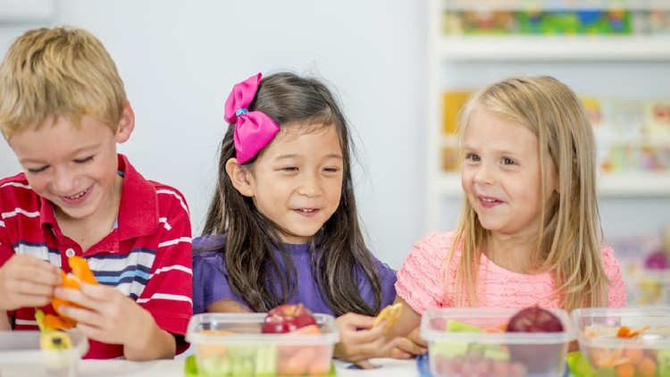 A multi-ethnic group of elementary age children are sitting together at a table eating their healthy snack full of apples, carrots, and celery.