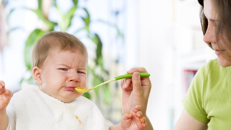 Crying baby boy refusing to eat food from spoon with hands dirty of vegetable puree.