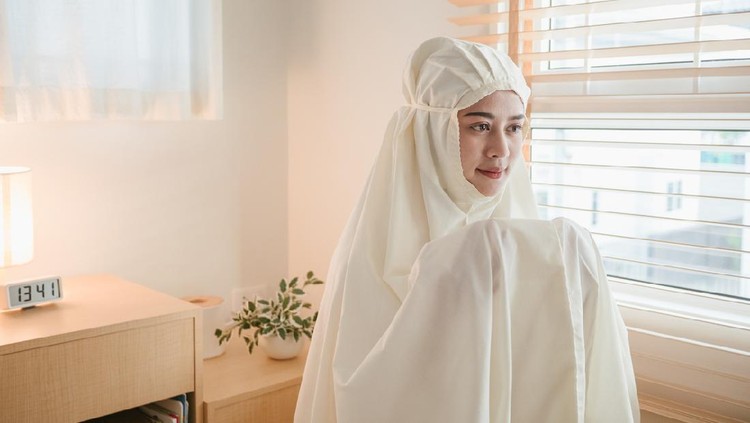 Young muslim woman in beige hijab and traditional clothes praying for Allah at home