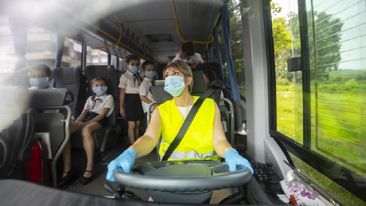 Young female bus driver driving he school bus, wearing a face mask. She is taking care that kids sit down.