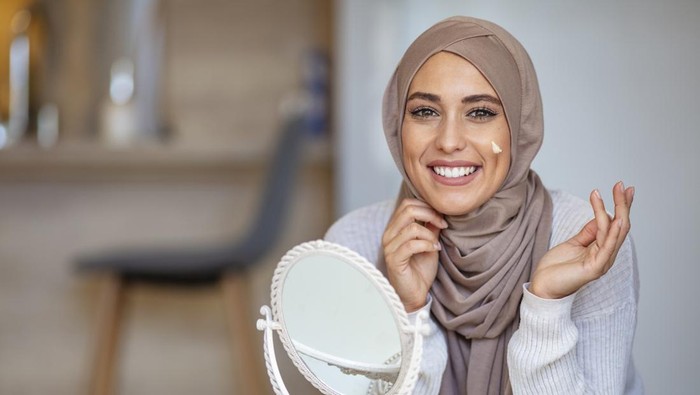 Muslim woman spreading cream over her face while looking in the mirror. Beauty treatment. Female putting on moisturizer on her facial. Muslim woman applying cream to face and looking to mirror