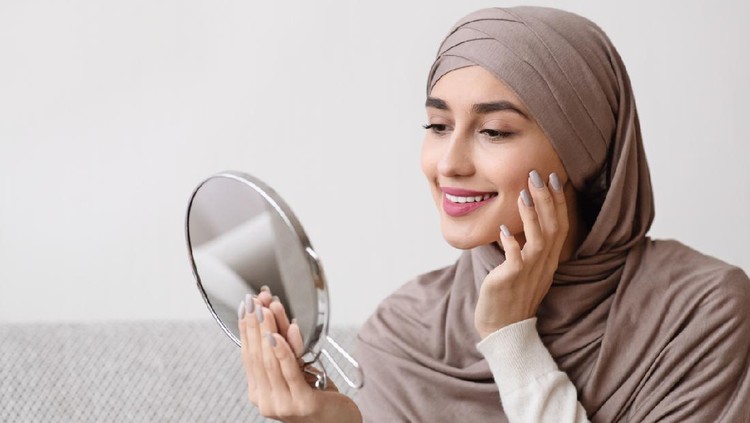 Beauty routine. Attractive muslim girl in hijab holding mirror and looking at her face with perfect soft skin, sitting on sofa at home, free space