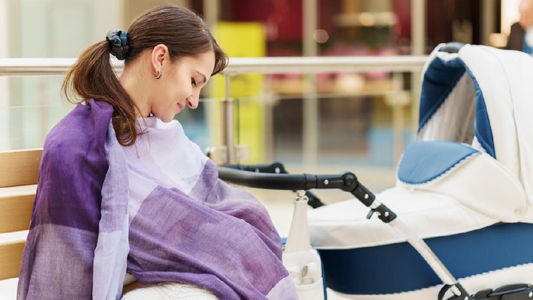 menyusui Young european woman with violet stole is breastfeeding her little child close to white baby carriage at public place shopping mall at day time