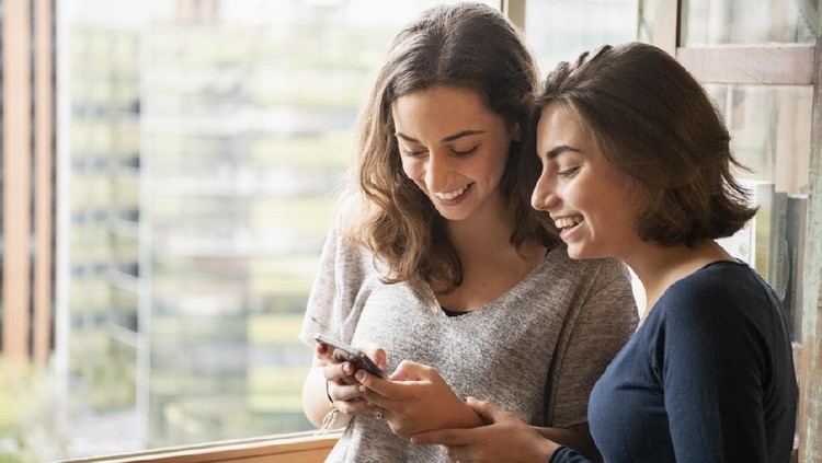 1068831124 Two sisters laughing and looking at messages on cell phone, online, social media, texting