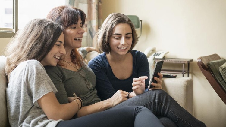 Brazilian mother laughing with two girls at home, with cell phone, on social media, smiling