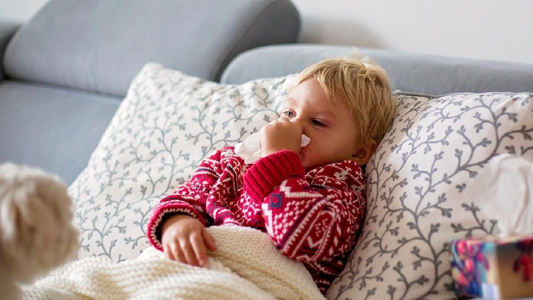 Sick child, toddler boy lying on the couch with his white puppy in living room with fever, resting at home