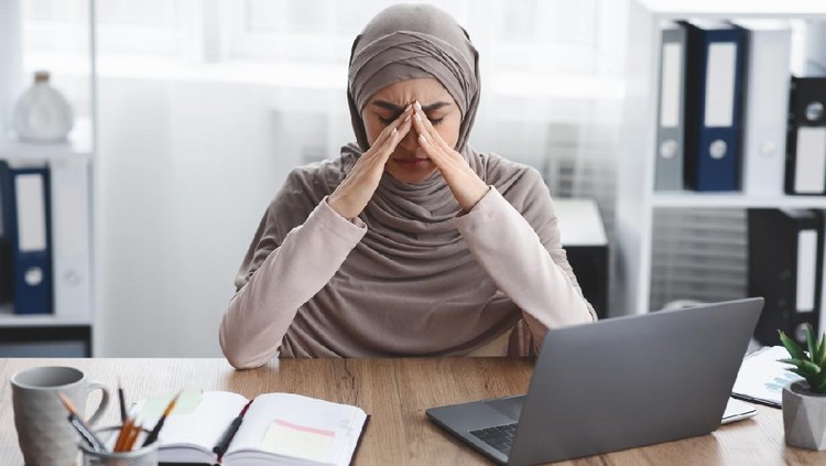 Exhausted arabic businesswoman having migraine after long time work in office, sitting at desk and touching her nose bridge, having bad day