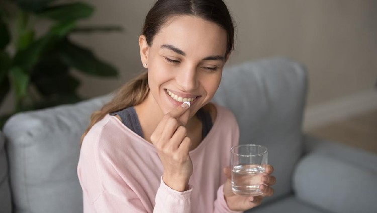 Young happy smiling mixed race woman sitting on a sofa at living room, holding glass of pure water, taking daily vitamin d, c complex, supplements for hair, skin and nail strengthen, healthcare concept.