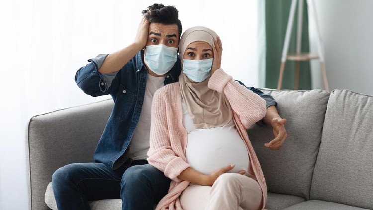 Pregnancy During Pandemic. Portrait Of Shocked Pregnant Muslim Couple In Protective Medical Face Masks Sitting On Couch At Home, Touching Heads In Horry, Afraid Of Coronavirus Outbreak, Free Space