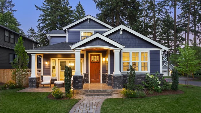 facade of home with manicured lawn, landscaping, and backdrop of trees and dark blue sky. Glowing interior lights create a welcoming mood.