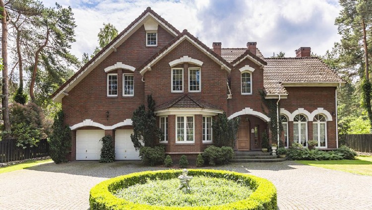 Front view of a red brick English style classic house with a steep roof, large windows and a circular driveway with a flowerbed decoration in the front.