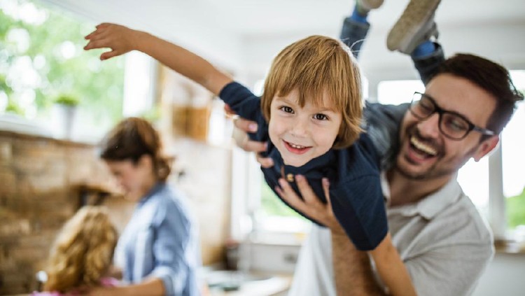 Happy little boy having fun with his father at home and looking at camera. There are people in the background.