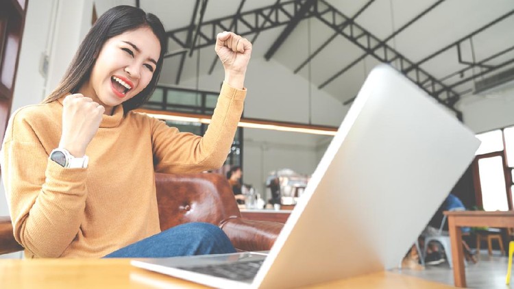 Young attractive asian woman looking at laptop computer feeling happy cheerful or excited expression success or win sitting on desk table in cafe coffee shop in wide close up view with soft tone.