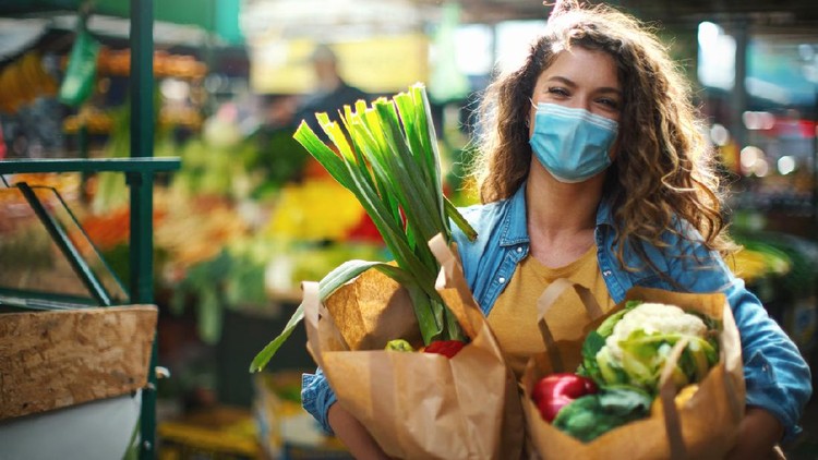 1282800704 Closeup side view of a late 20's woman going grocery shopping during coronavirus pandemic. She's at a farmer's market with bags full of fresh fruit and vegetables, smiling and looking at the camera,