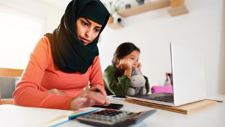 Young Muslim mother calculating while her daughter is sitting next to her looking at the laptop screen during coronavirus quarantine.