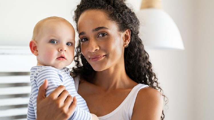 Smiling african mother holding adopted baby at home. Portrait of proud young woman rocking infant while looking at camera. Beautiful mixed race babysitter carrying adorable little child at home.