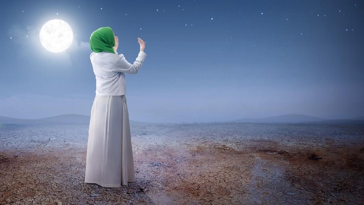 Rear view of asian muslim woman in veil raised hands and praying on the sand dune with moon and night scene background