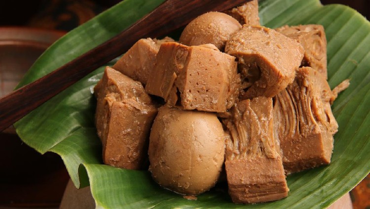 Close-up view of Gudeg, the signature dish of jackfruit stew from Jogjakarta. Chunks of jack fruit and eggs stewed with coconut milk, palm sugar, and teak leaves; commonly done in a traditional earthenware cooking jar known as kendil. Here shown the gudeg is arranged on a banana leaf that is placed on a kendil. A pair of wooden tongs is tucked on side further back.