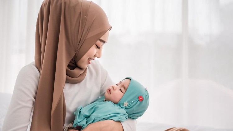 Happy muslim mother holding adorable little baby daughter wearing hijab in her arms on white bed in bedroom. Arab young mom wearing a head scarf, and she's sitting on the bed while holding her baby.